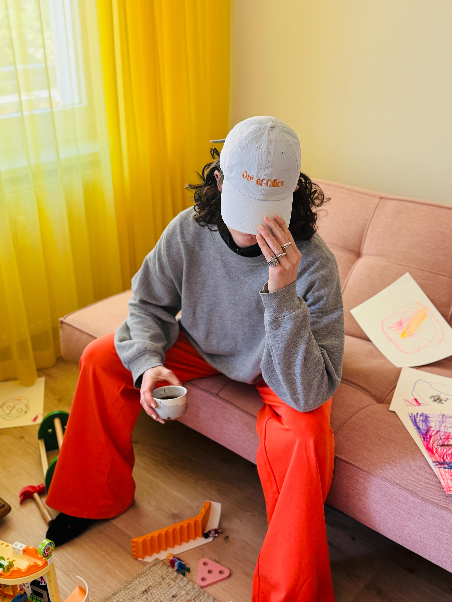 Caecilia Out Of Office Eine Frau in einem grauen Sweatshirt und einer orangefarbenen weiten MIUMI-Hose sitzt auf einer blassrosa Couch. Sie hält eine Tasse in der Hand und bedeckt ihr Gesicht mit einer Mütze, auf der „Out Of Office“ steht. Verspielte, leicht chaotische Atmosphär
