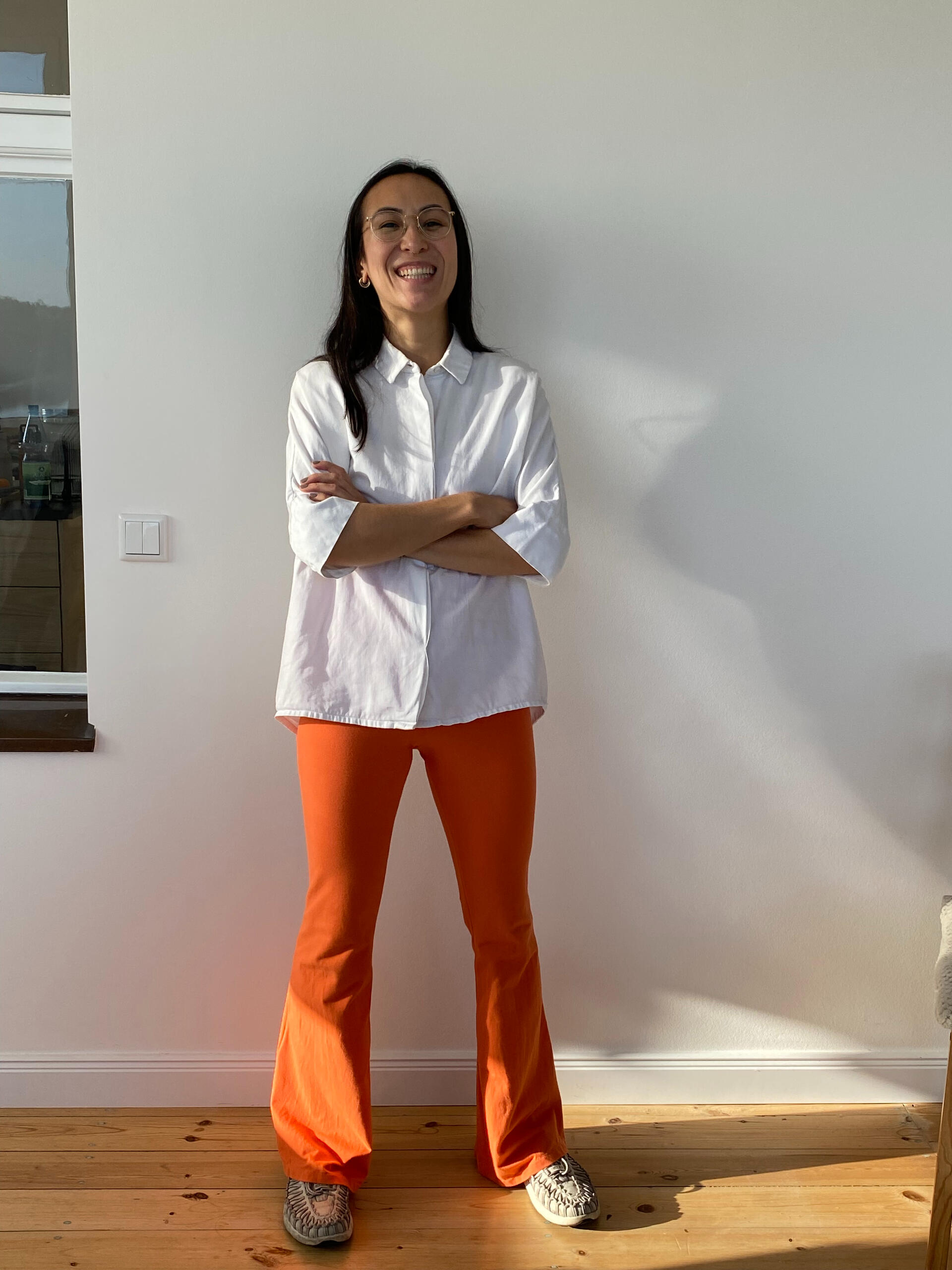Breathwork Lehrerin Kim Gerlach in MIUMI Schlaghose in Orange. Eine Frau steht lächelnd mit verschränkten Armen da. Sie trägt ein weißes Button-up-Hemd und eine leuchtend orangefarbene MIUMI-Schlaghose. Der Schauplatz ist ein Raum mit hellem Holzfußboden.