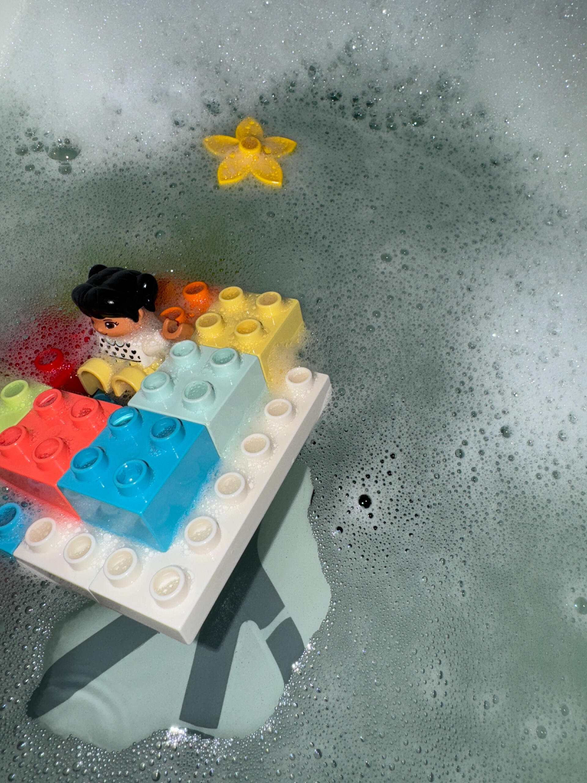 LEGO Boot Eine Spielzeugfigur sitzt in einem Boot aus bunten LEGO Steinen inmitten von Seifenblasen in einer Badewanne. In der Nähe schwimmt ein gelber Spielzeugstern.
