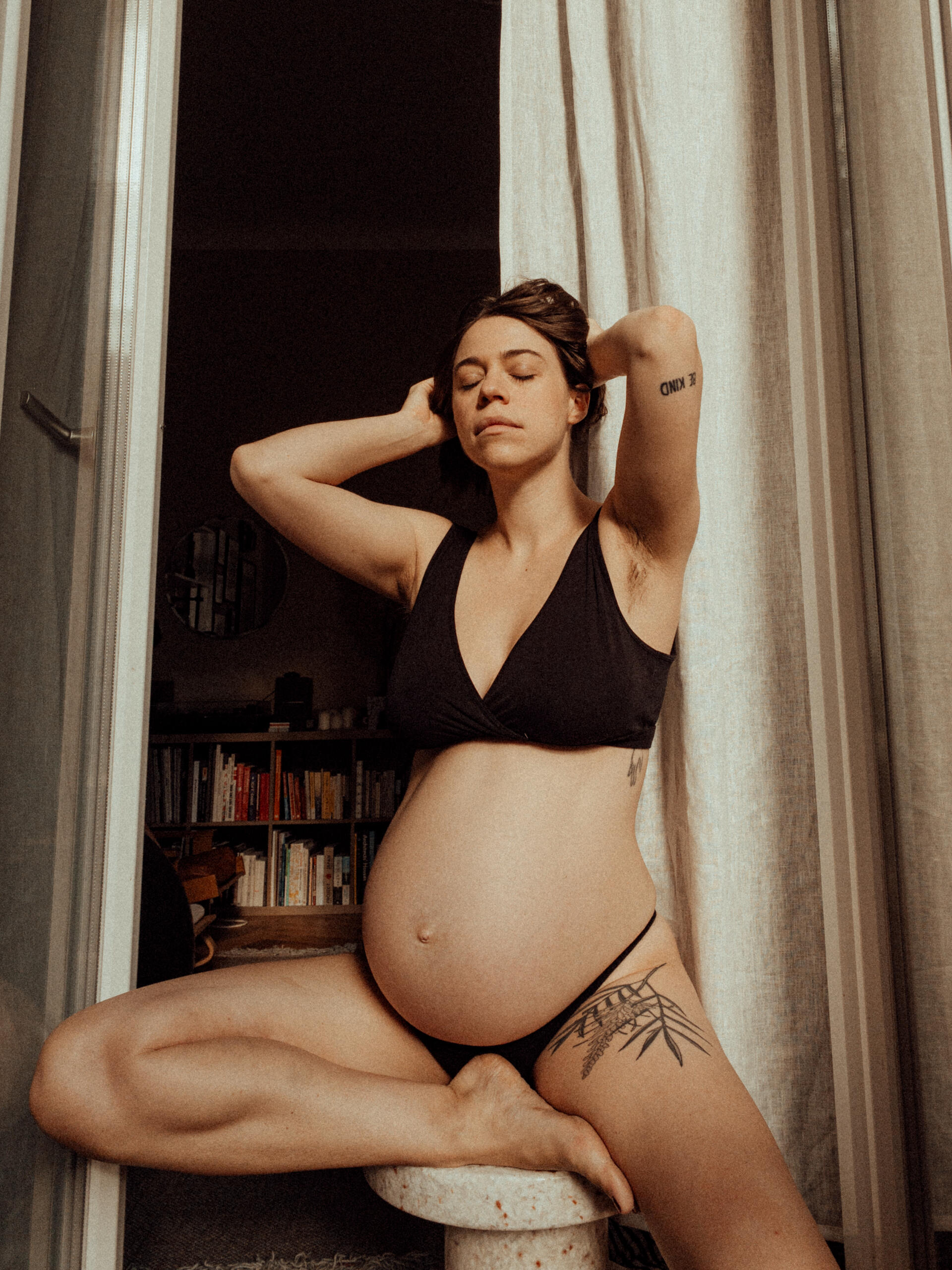 Pregnant Kat in full bloom A pregnant woman, wearing only underwear, sits on a chair in front of a white curtain. Her eyes are closed – a quiet moment, caught in warm, intimate light.