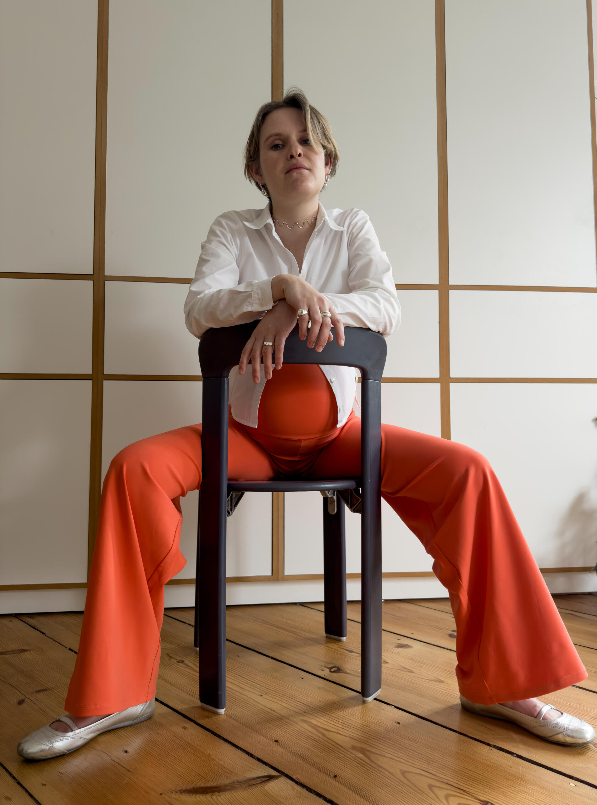 Sonja in MIUMI wide leg pants in orange. Pregnant woman in a white shirt and bright orange MIUMI maternity pants sitting on a chair backwards.