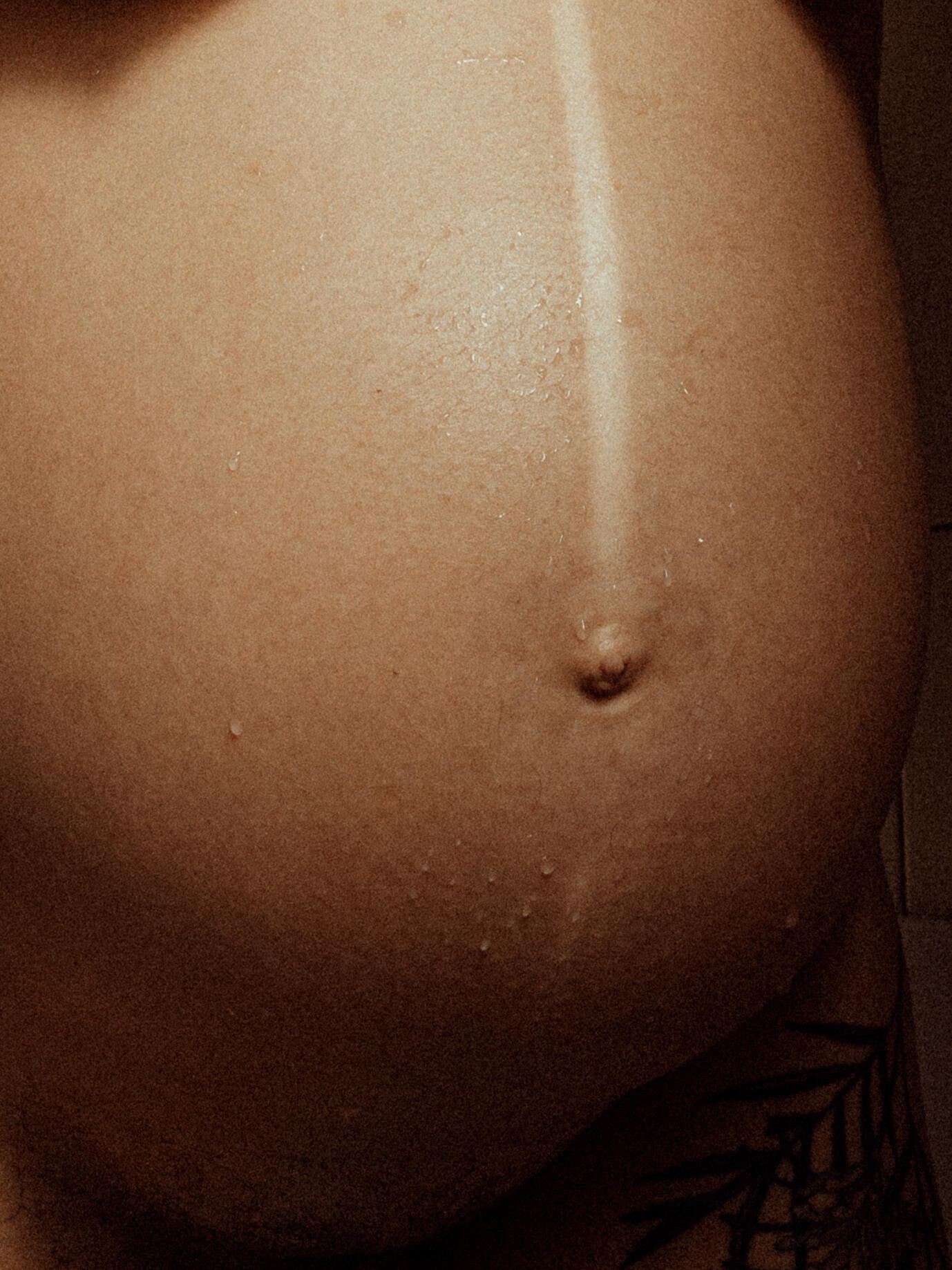 Kat's baby bump Close-up of a pregnant belly with a prominent linea nigra and droplets of water. A tattoo peeks from the lower right, conveying intimacy and strength.