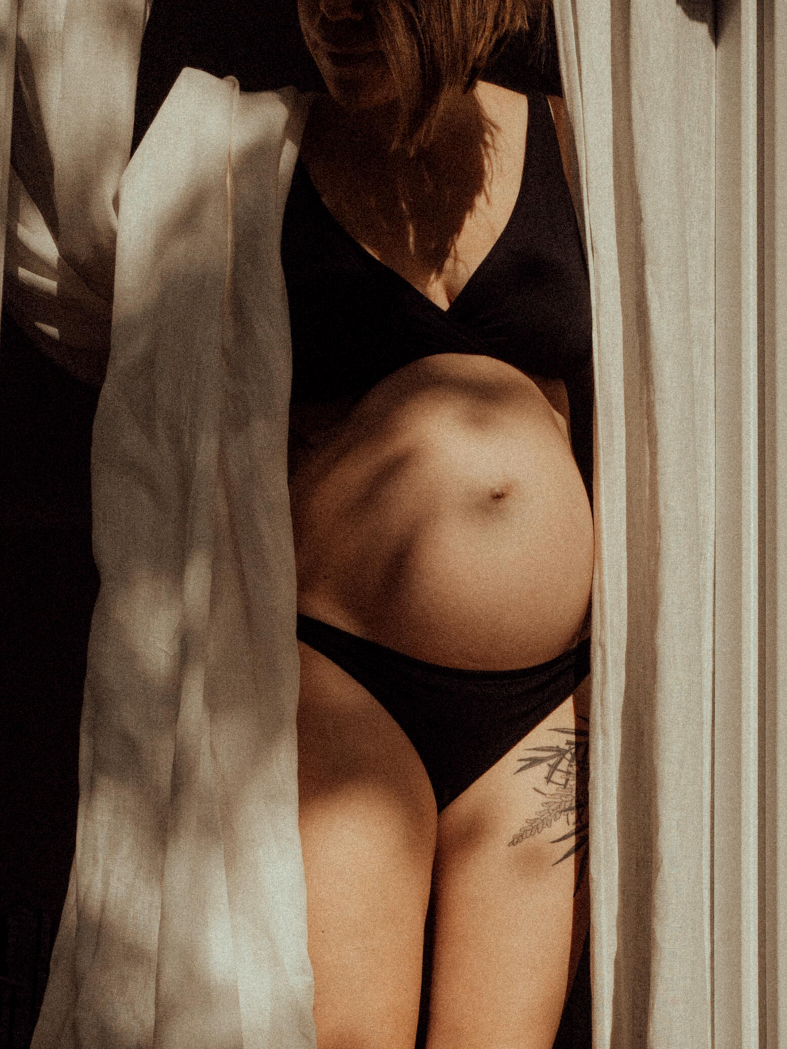 Kat In Her Own Light Pregnant person in black underwear, partially covered by white curtains, stands in warm light, evoking a serene and intimate atmosphere.
