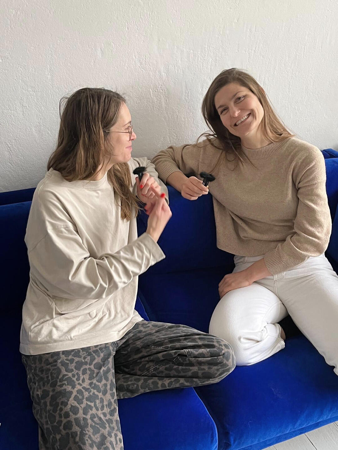 Hanna Stutzki visits MIUMI Two women (Antonia Pahlke von MIUMI & Hanna Stutzki) are sitting on a blue sofa, smiling and holding black microphone clips. One wears a beige sweater, the other a cream top and patterned pants.