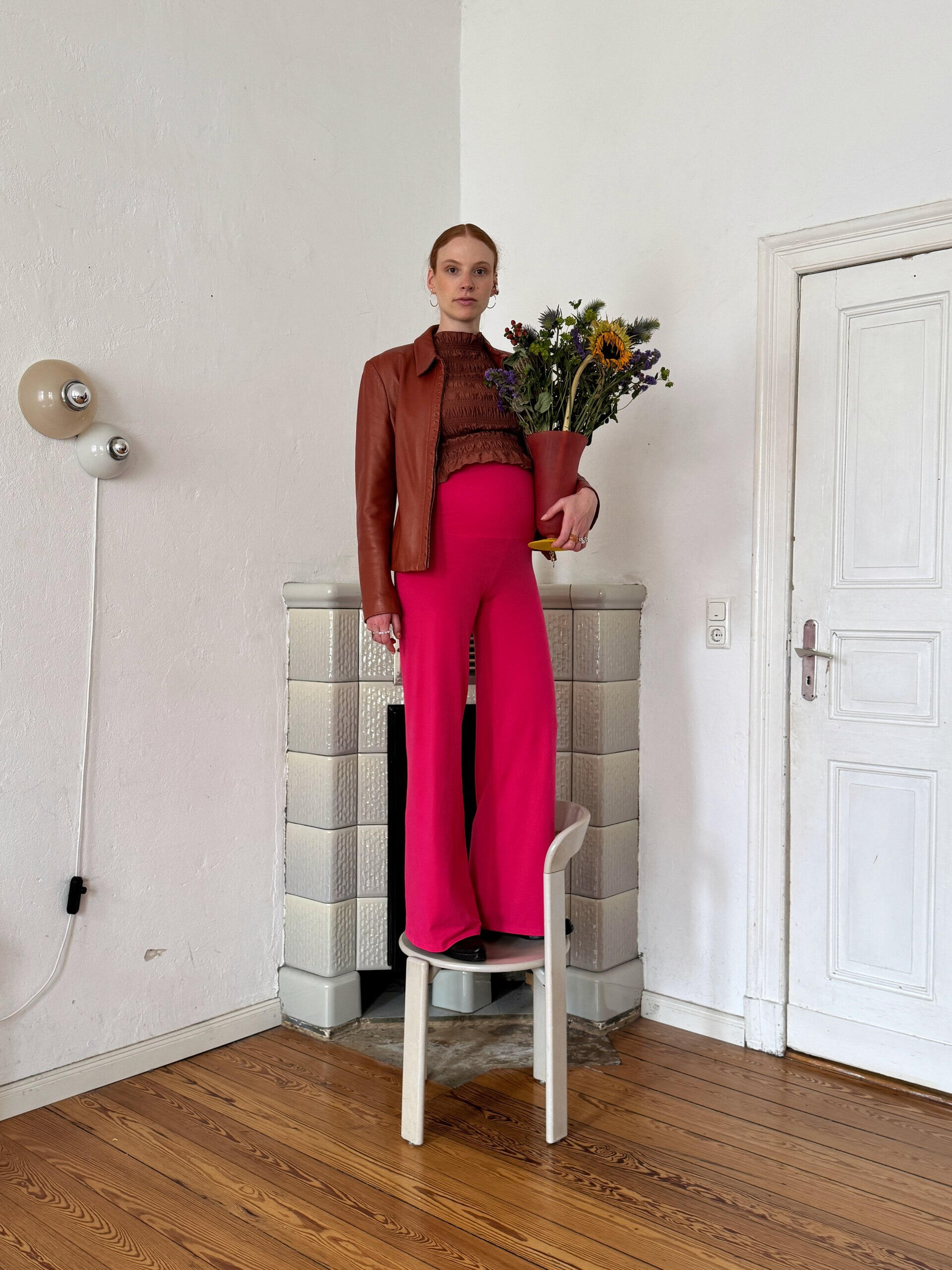 Katharina Bellinger loves bold design A pregnant woman in a brown jacket and vibrant pink MIUMI pants stands confidently on a chair, holding a vase full of dried flowers, against a minimal indoor setting.
