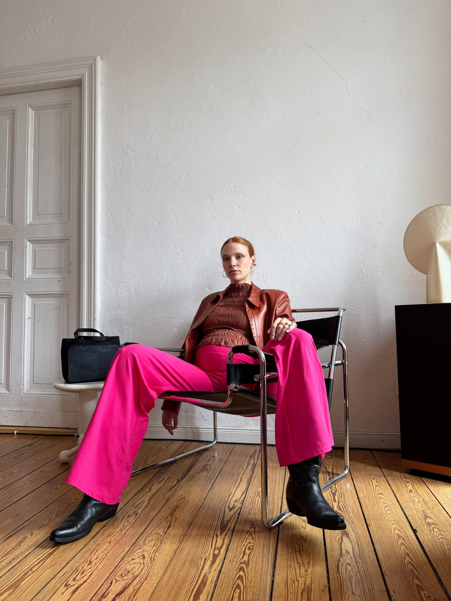 Katharina Bellinger in her curated home Pregnant woman seated in a Wassily lounge chair wearing a brown jacket, pink MIUMI pants, and black cowboy boots. She exudes a confident, relaxed pose in a stylish room.